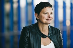 A dark-haired woman, Amazon's disaster relief head Bettina Stix, is dressed in a black leather jacket looks to the right and smiling. There is a blue background. 