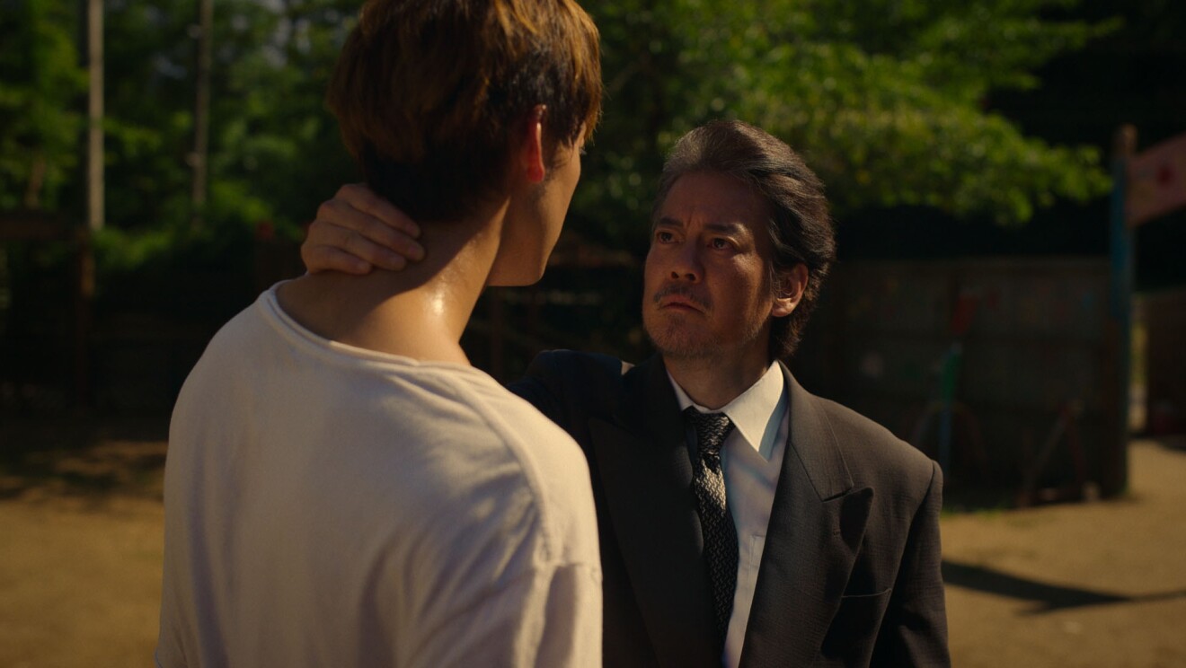 白いTシャツの男性の首を右手でつかみ、その男性を見つめているダークシャツの白髪交じりの男性