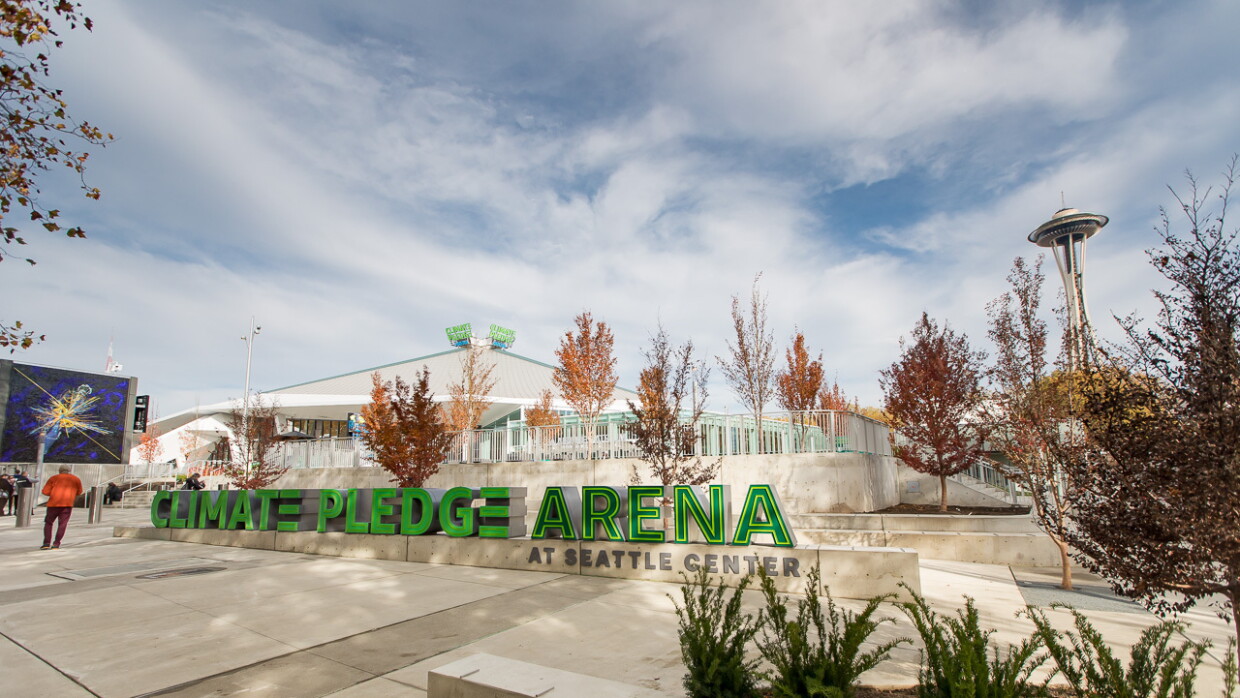 Climate Pledge Arena a Seattle, EE.UU. Parte exterior del estadio cubierto. Aparece en letras grandes y de color verde Climate Pledge Arena at Seattle Center en primer plano. De fondo el estadio. el suelo es de mármol y hay árboles de color marrón. 