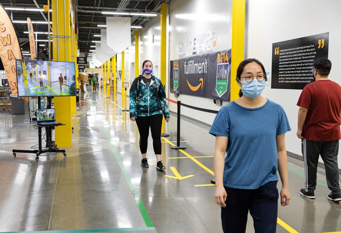 Amazon associates wearing face masks walk along a corridor of a fulfillment center. Behind them, a screen shows visual social distance assistance.