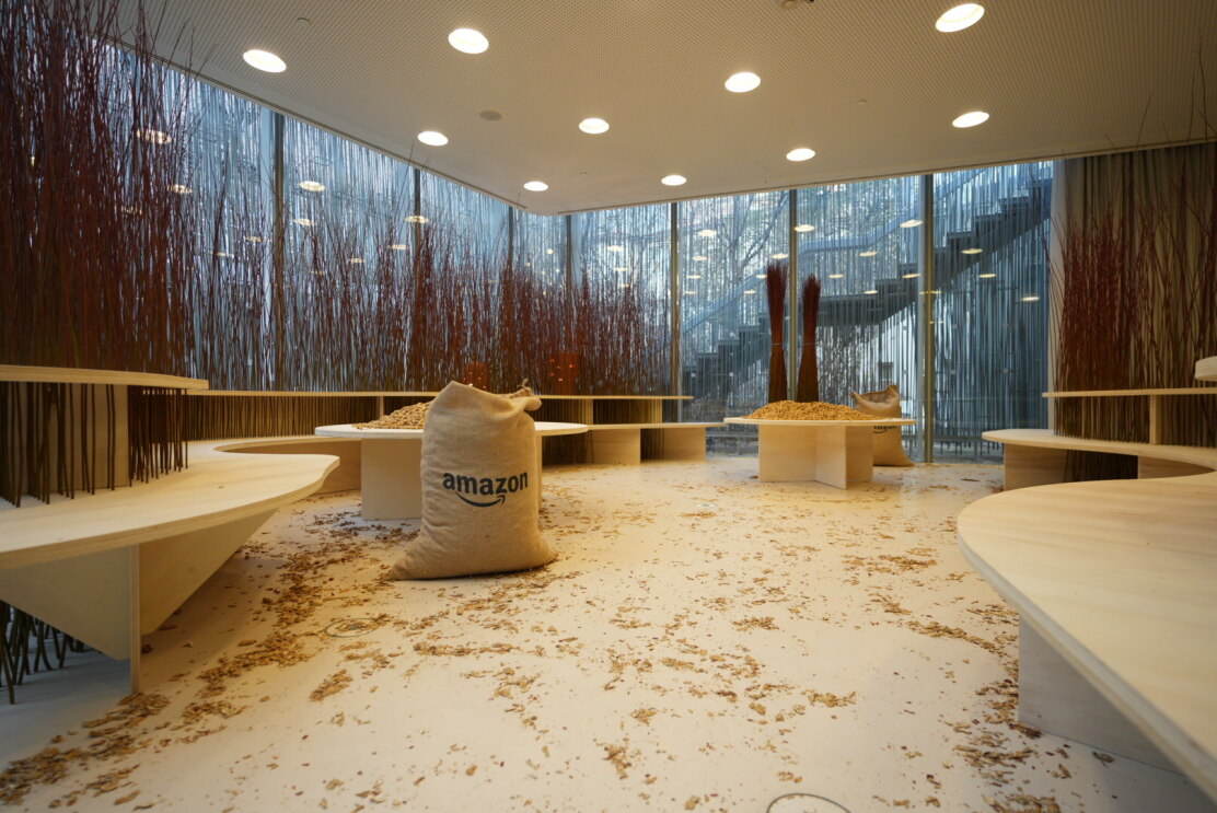 Un espacio sin gente, con el suelo blanco y paredes de cristal. Sobre el suelo hay cáscaras de cacahuete tiradas. A los lados, dos bancos con formas curvas, de madera. Detrás de los bancos, varas de mimbre en posición vertical. La luz es cálida y hay un saco de rafia con el logo de Amazon. 