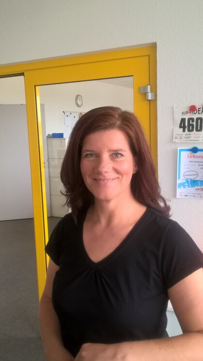 Eine Frau mit langen braunen Haaren und scharzem T-Shirt steht lächelnd an einem Bürotresen.