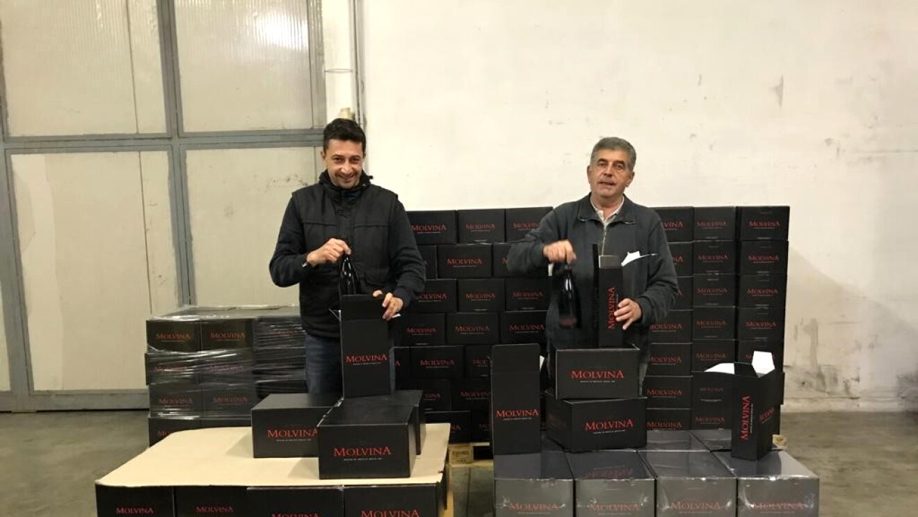 Due uomini in un capannone di fronte a due pallet con delle casse di vino in scatole nere. 