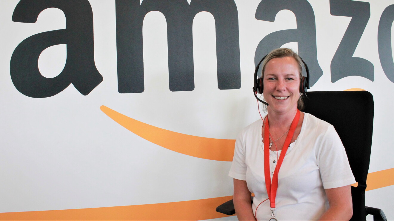 Eine Amazon Kundenservice-Mitarbeiterin mit Kopfhörer vor einer Wand mit Amazon Schriftzug.