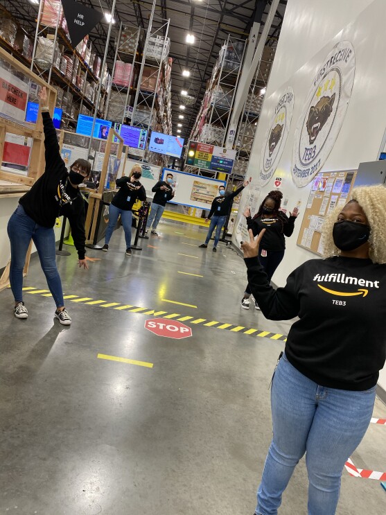 Six Amazon employees stand in an Amazon fulfillment center, preparing for Peak. All are wearing Fulfillment shirts, face masks, and are socially distanced.