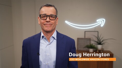 Doug Herrington wears a navy suit and stands in front of a lit up Amazon smile logo hanging on the wall.