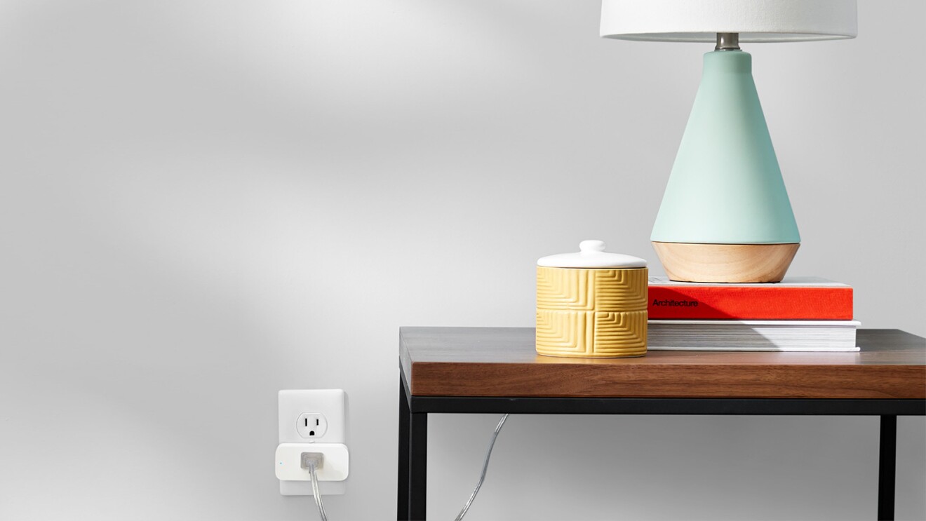 An image of a lamp sitting on a wooden table on top of two books and with a yellow container next to it. The lamp is plugged to a wired smart switch on the left.