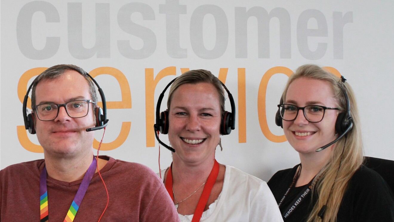3 Amazon Kundenservice-Mitarbeiter:innen mit Kopfhörern. Im Hintergrund kann man auf der Wand Amazon Customer Service lesen.