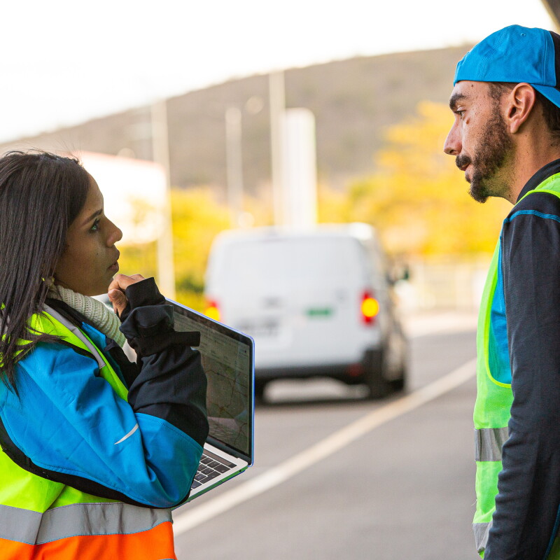 Ivette Duran, CEO de Cainaima Logistics, de pie con un ordenador entre las manos y hablando con un conductor de reparto de paquetes. Están los dos en la calle y de fondo hay dos furgonetas blancas. Ella es morena y tiene el pelo largo y negro. Lleva un jersey de color blanco y de cuello alto, una chaqueta azul y un chaleco fluorescente. Ell es más alto, con barba y bigote y lleva una chaqueta de color azul y gris y un chaleco fluorescente. Los dos están hablando cara a cara. 