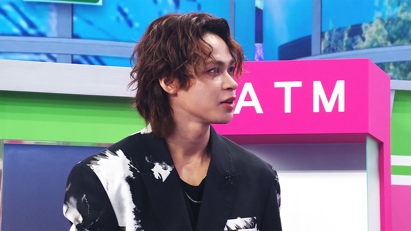 一部が黄緑やピンクに塗られたセットを背景に、黒いジャケット姿の男性の上半身。顔だけ右に向いてほぼ横向きになっている。背後のピンクに塗られた部分に白字で「ATM」。