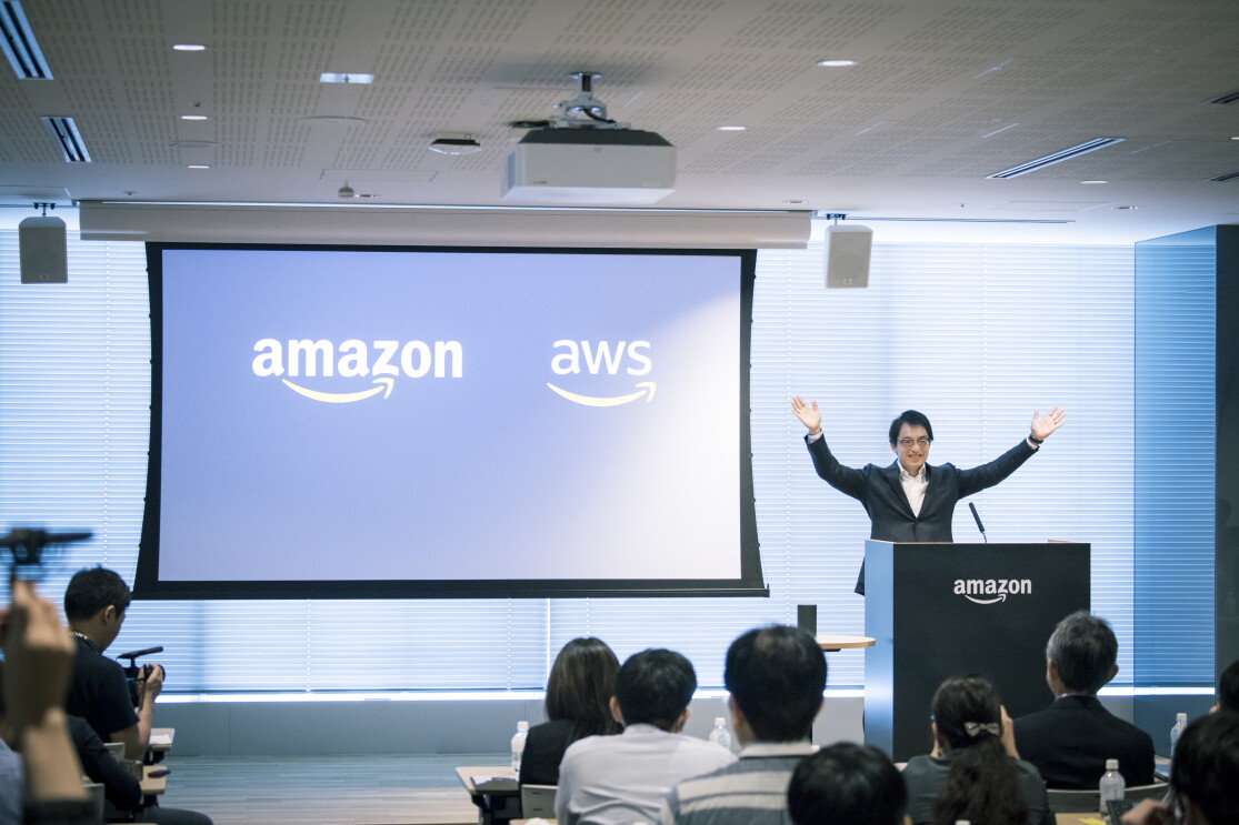 オフィスの会議室に集まった人々。壁の前にスクリーンがあり、AmazonとAWSのロゴが投影されている。右側の登壇者が両腕を挙げている。