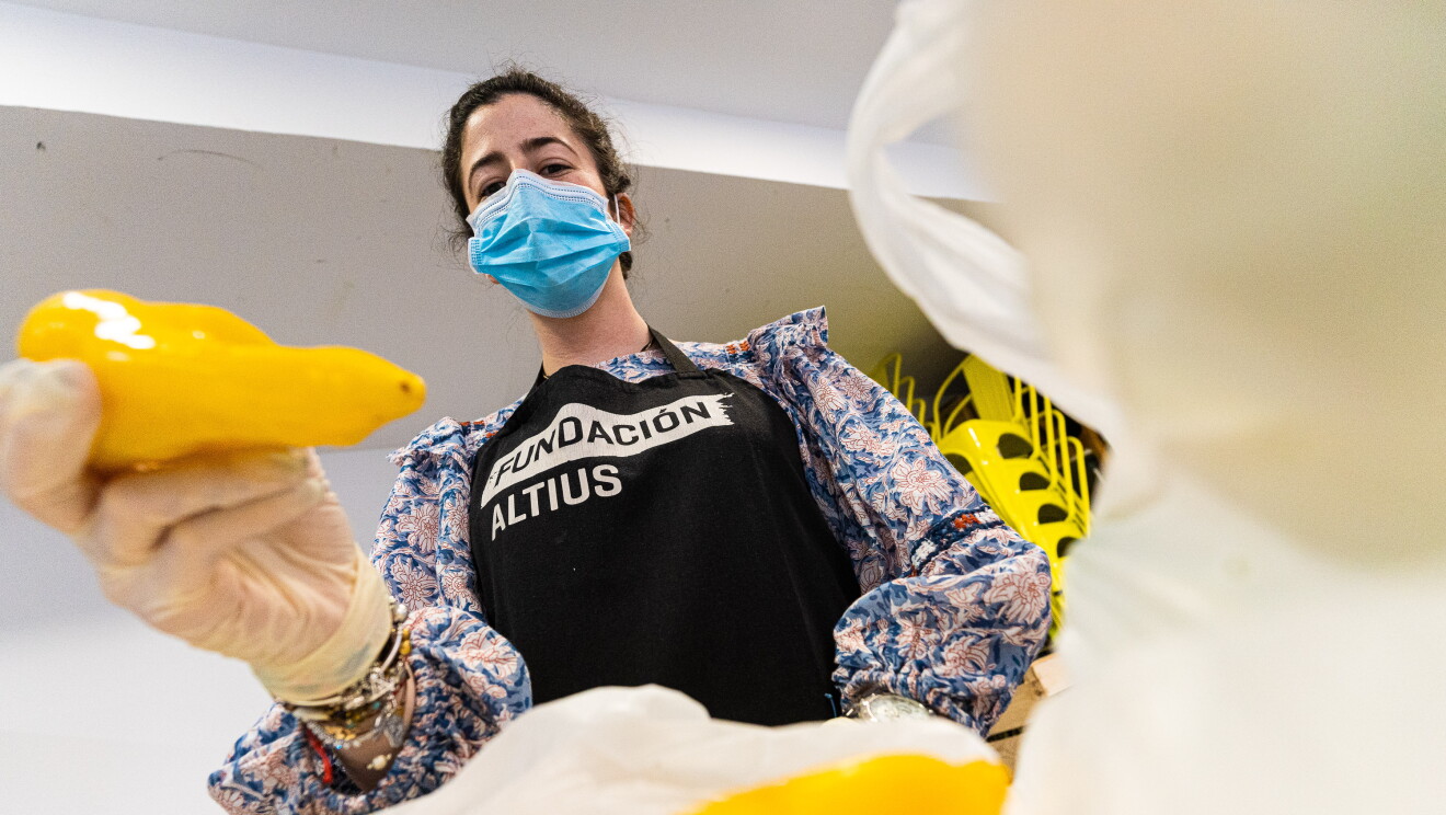 Una voluntaria de la Fundación Altius llenando bolsas de comida para familias vulnerables. Ella vista una camisa de flores de manga larga, un delantal negro con las letras en blanco de FUndación Altius, LLeva el pelo recogido, una mascarilla y guantes blancos. En la mano pimiento de color amarillo que está a punto de introducirlo en la  bolsa. La perspectiva del lector es como si estuviera dentro de la bolsa de comida. 