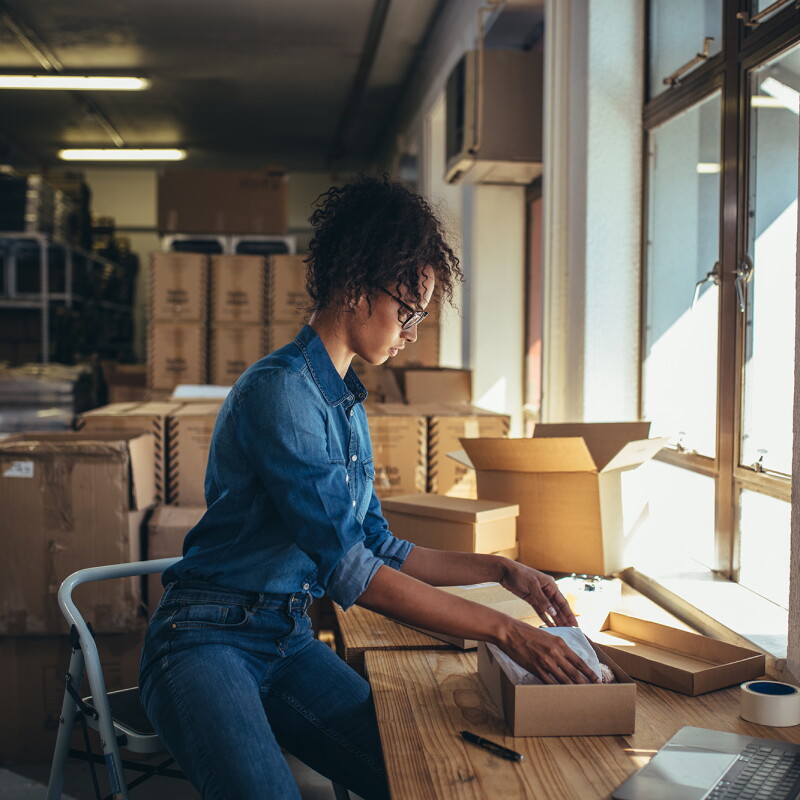 A woman sits on a step stool as she packages a product in a kraft box. 