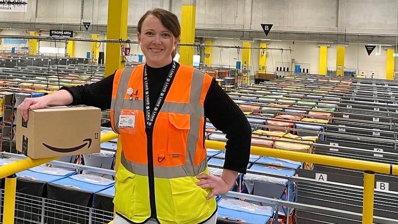 Vera Steitz im Amazon Verteilzentrum Rostock: Sie steht mit einem Päckchen in der Hand und blickt lächelnd in die Kamera. Im Hintergrund sieht man mit Blick von oben die Regale im Verteilzentrum.