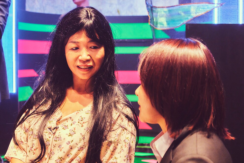 黒地にピンクと黄緑の横縞が入ったセットで、話し合う2人の女性のアップ。左側の女性は長い黒髪のかつらに小花柄の服、右側の女性は黒いスーツを着用で横顔だけ見えている。