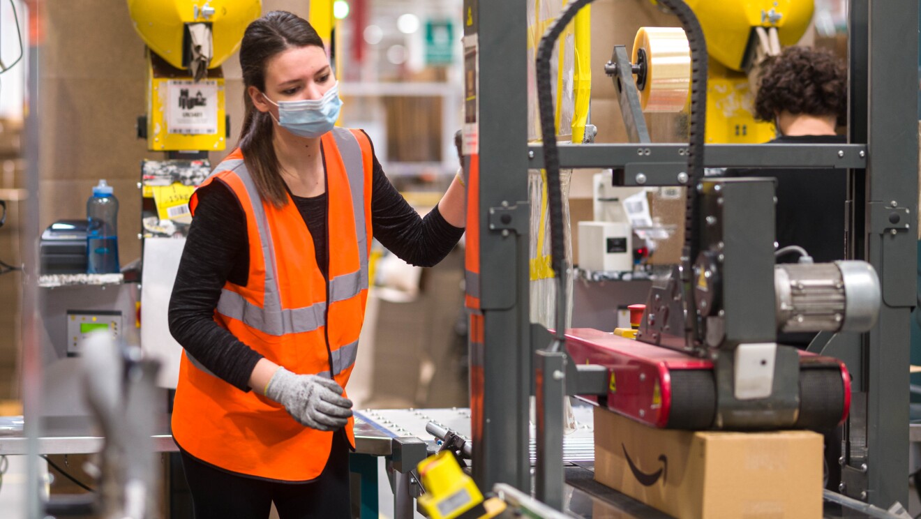 Inquadratura di una dipendente di un magazzino Amazon che sta maneggiando un attrezzo per apporre etichette sui pacchi. In basso a destra si intravede un pacco sotto la pressa che incolla le etichette.
