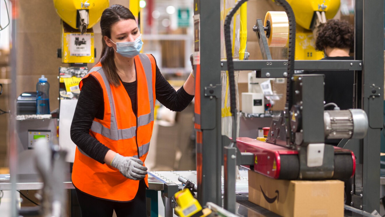 Inquadratura di una dipendente di un magazzino Amazon che sta maneggiando un attrezzo per apporre etichette sui pacchi. In basso a destra si intravede un pacco sotto la pressa che incolla le etichette. 