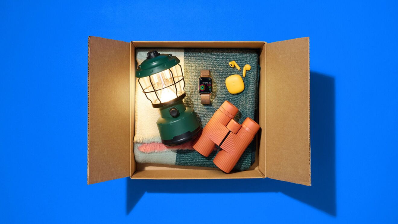 Open cardboard box containing camping gear including green lantern, orange binoculars, smartwatch, and yellow water bottle on folded denim fabric