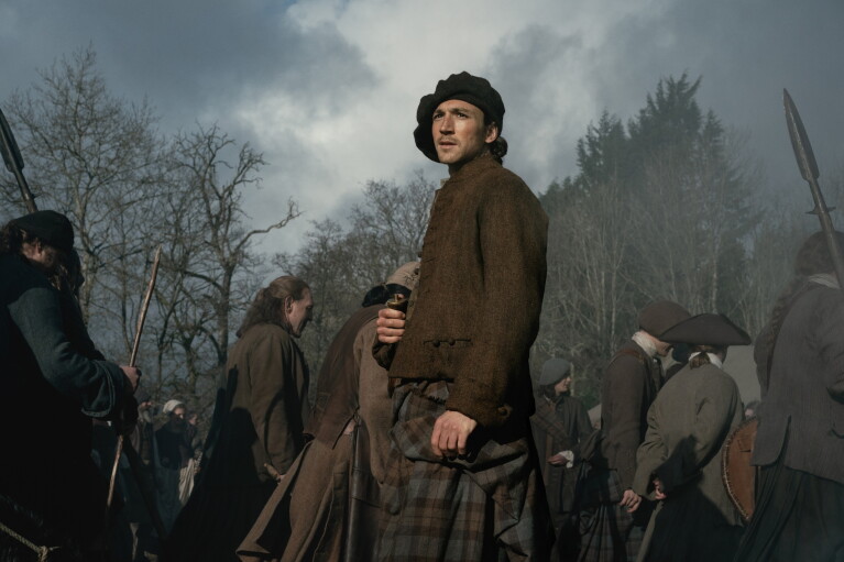 Man in traditional Scottish attire stands amid gathering crowd in misty forest