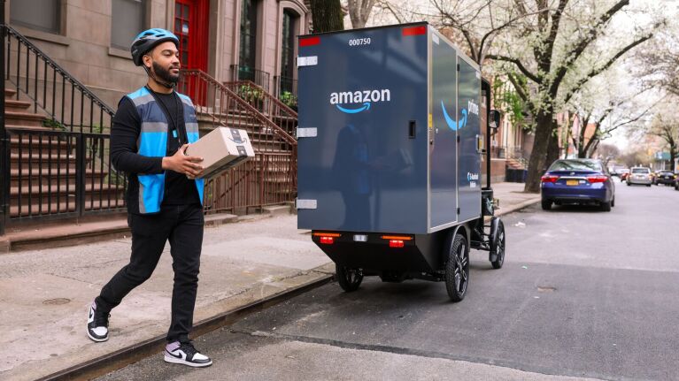 Amazon delivery worker with package next to autonomous vehicle on city street