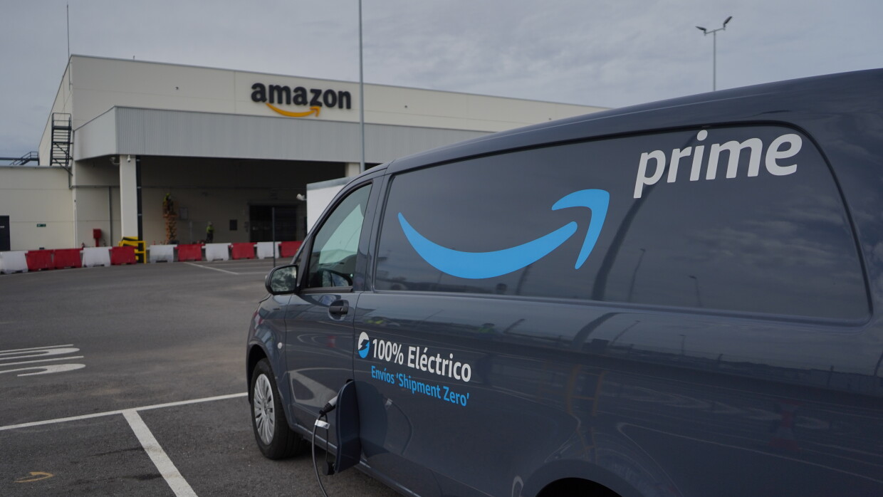 Una Mercedes Benz-Vans en un aparcamiento. Se ve el logo Prime de Amazon y el texto en la puerta 100% Eléctrico Envíos 'Shipment Zero'. En primer plano se ve un cargados de batería que está subministrando electricidad a la furgoneta de al lado que solo se ve un poco su parte trasera. 