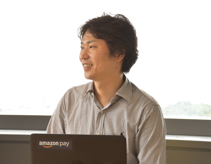 窓を背に話すグレーのシャツを着た男性。窓の外はほとんど白い空で、遠くに緑が見える。体の前に黒いノートパソコンを開いている。ノートパソコンの蓋に「amazon pay」の文字。