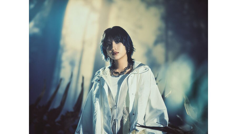 森のような屋外で木漏れ日を受けて立つ人物。白い衣装を着用、重そうなチェーンのネックレスを首にかけている。