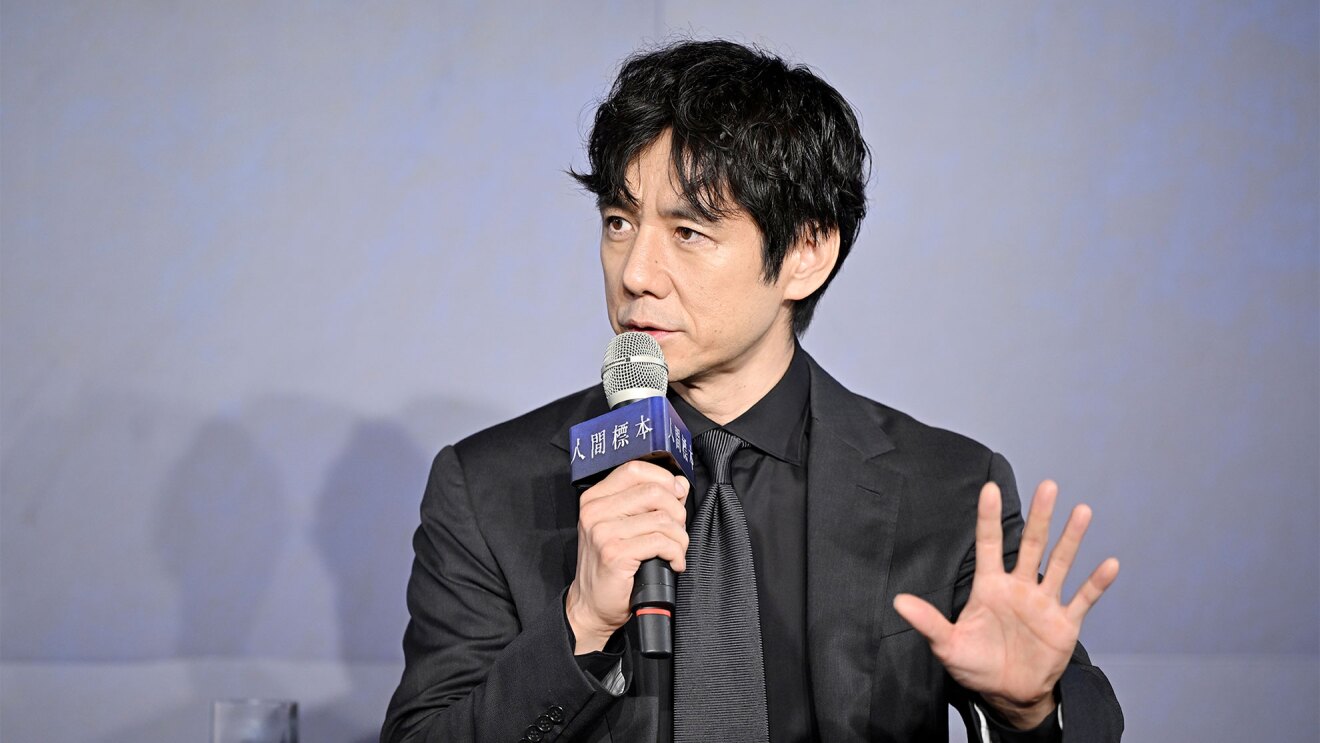 グレーの背景に、スーツ、シャツ、ネクタイすべて黒を着用している男性。右手を開いて前に出し、左手に持ったマイクには「人間標本」と書かれたプレートが付いている