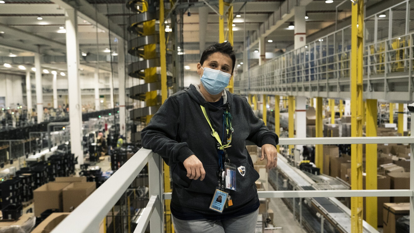 Una donna con i capelli corti indossa una mascherina chirurgica sul volto e poggia i gomiti su una balaustra alle sue spalle. Sullo sfondo di vede l'interno di un magazzino Amazon. 