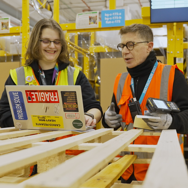 A young woman and a man with yellow and orange vests are looking at something on a laptop screen inside a fulfillment center