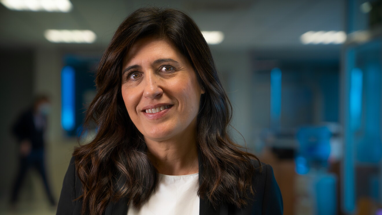 Ruth Alonso con una americana negra y un jersey gris. Sonriente mira a cámara sin mascarilla. Tiene el pelo negro por debajo de los hombros. De fondo desenfocado lo que podría ser una oficina. 