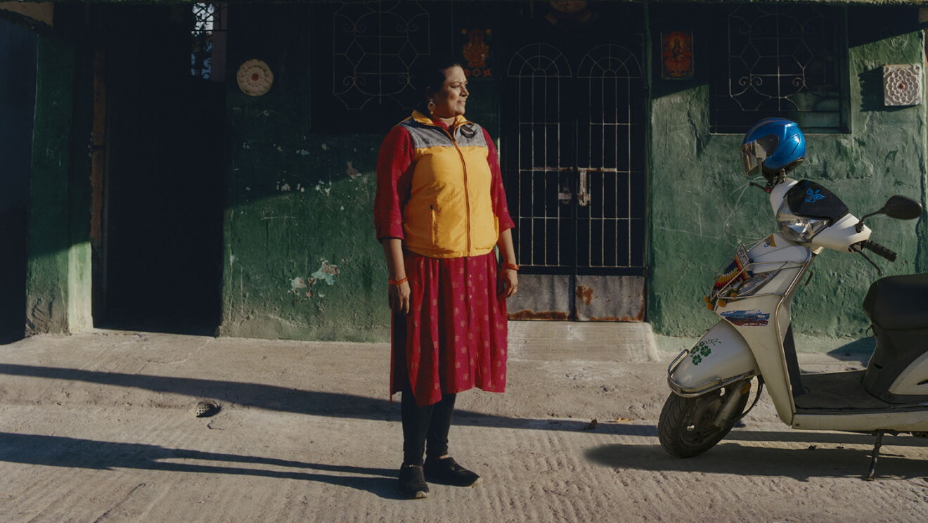 Una mujer india de pie mirando su moto de color blanco con un casco azul en el manillar. La mujer va vestida con un chaleco amarillo de Amazon y un vestido de color rojo. De fondo una casa con la pared verde. 