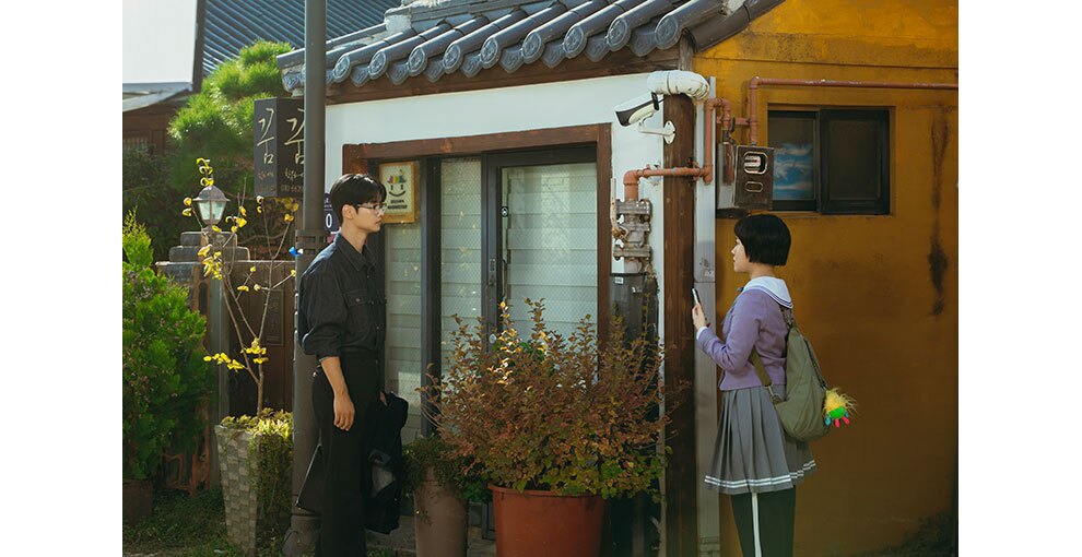 日中の屋外で、向かい合って見つめ合う男性と制服の少女。背後にはハングル文字の看板がかかった建物。