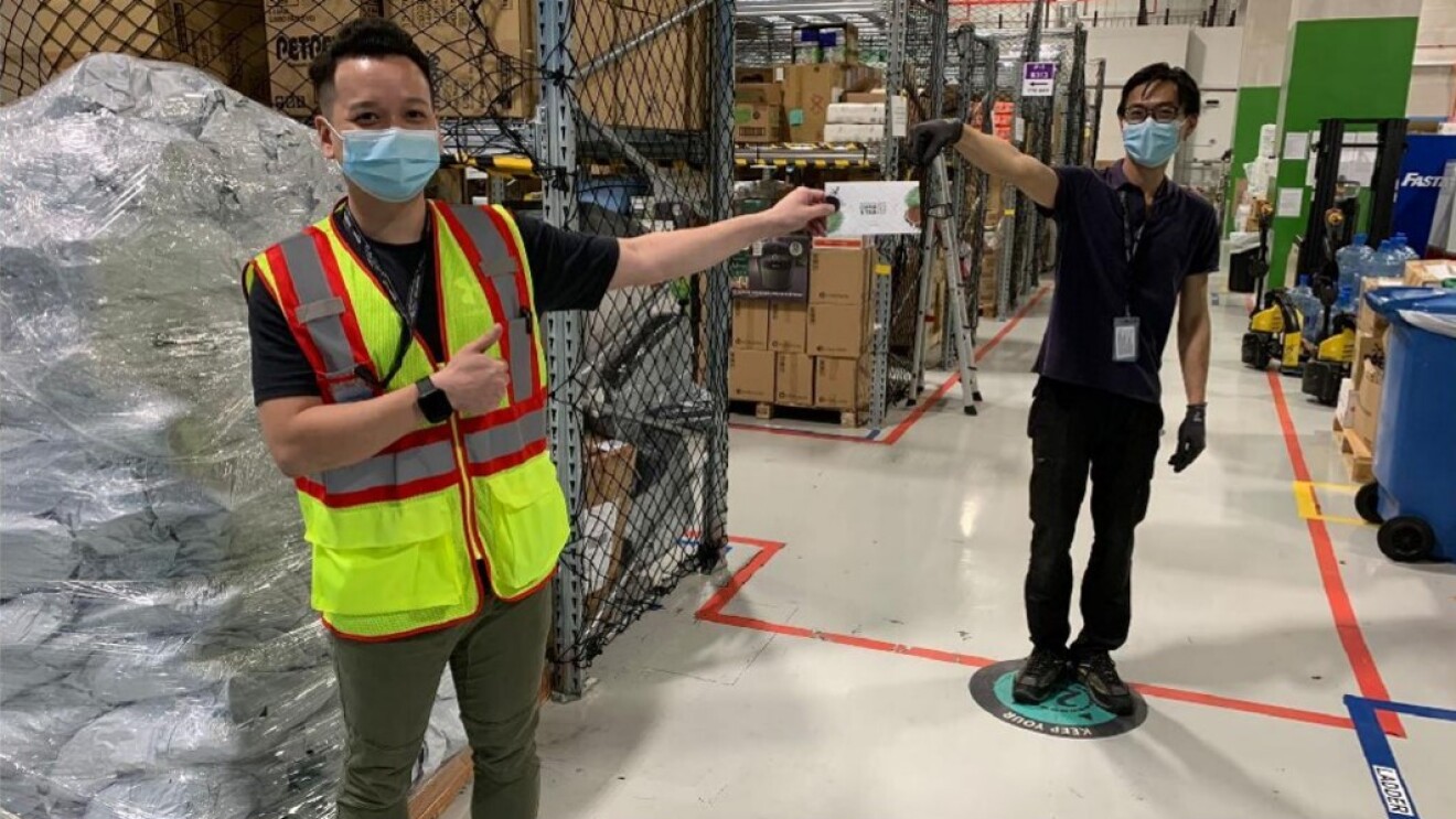 Two men can be seen standing at a distance of two meters from each other. One person is handing over an envelope to the other. Both men are wearing masks. The man on the left side is wearing a safety vest