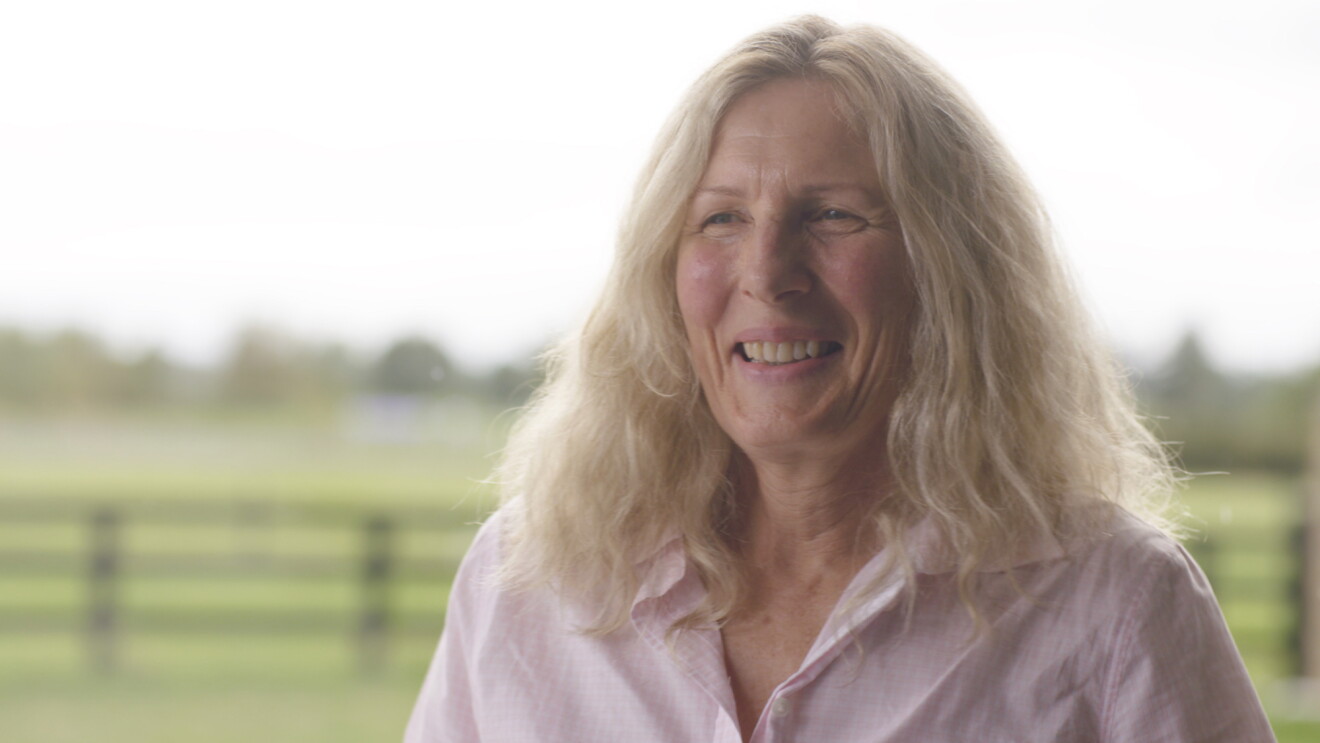 A woman smiles at the camera, behind her a rural setting can be seen.