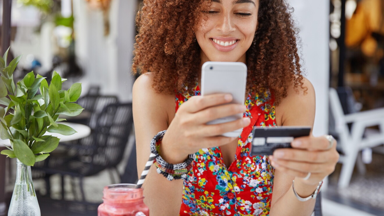 Foto de uma mulher, sorrindo, segurando um celular e um cartão de crédito faz compras online