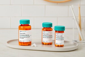 Three Amazon Pharmacy pill bottles sit on a white tray next to a glass with two toohbrushes in it.