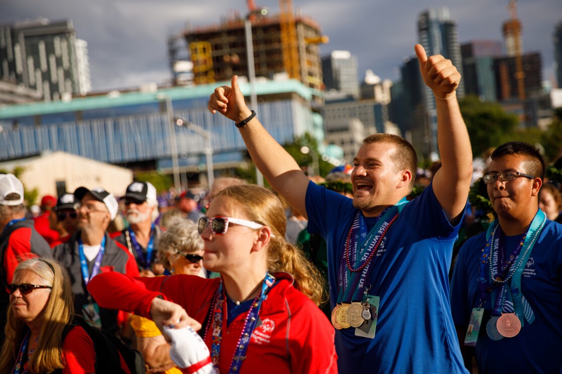 Special Olympics athletes and closing ceremony attendees celebrate. Many people looking toward the stage, with a male athlete giving thumbs up while smiling.
