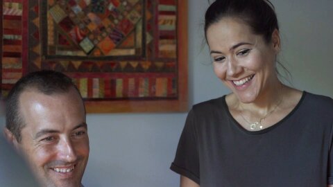 El escritor Mario Villén y su esposa, los dos sonriendo, miran la pantalla del ordenador. En la pared hay una tela colgada y la esposa luce un jersey gris con un collar de oro.     
