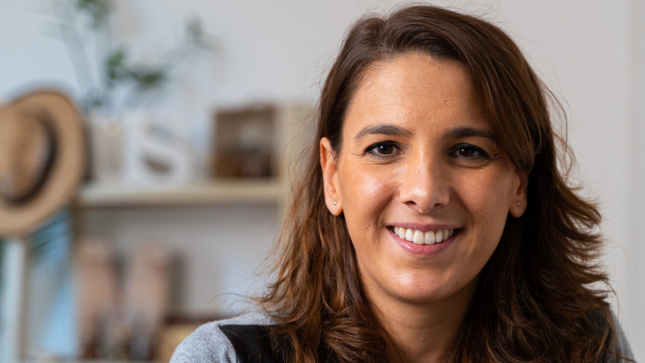 María Garvía. Mirando a cámara con una sonrisa y el pelo largo de color marrón pero escalonado. Pendientes y flequillo. Jersey gris y negro. De fondo, desenfocada, una estantería de una casa con fotos, plantas y un gorro de mimbre. 