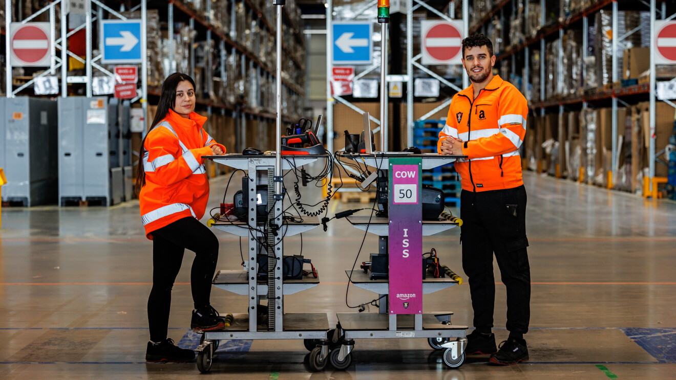 Miriam y Mario apoyados cada uno en una mesa alta transportable de Amazon. Los dos apoyan los brazo en la mesa. La mesa de él tiene un ordenador, la de ella tiene un ratón y un lector de código de barras. Los dos van con una chaqueta naranja que simula un chaleco de seguridad. De fondo el pasillo de un centro logístico. Miriam lleva el pelo largo por la cintura y es de color castaño. Él tiene el pelo corto pero la parte superior la lleva un poco más larga y la tiene rizada. Tiene los ojos claro, barba de dos días y un piercing en el labio inferior. De fondo las estanterías de un centro logístico.
