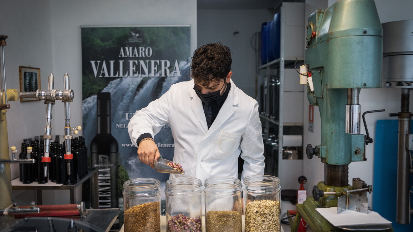 Maurizio Cappelletti all'interno del sito di produzione dell'amaro Vallenera. Indossa un camice e una mascherina e davanti a sè ha dei vasi in vetro con le erbe e le spezie con cui produce i liquori. Sullo sfondo un poster con il logo Amaro Vallenera