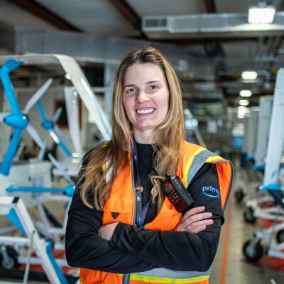 An image of Christina Carter smiling for a portrait at the Amazon drone facility in Texas. There are several rows of drones behind her.