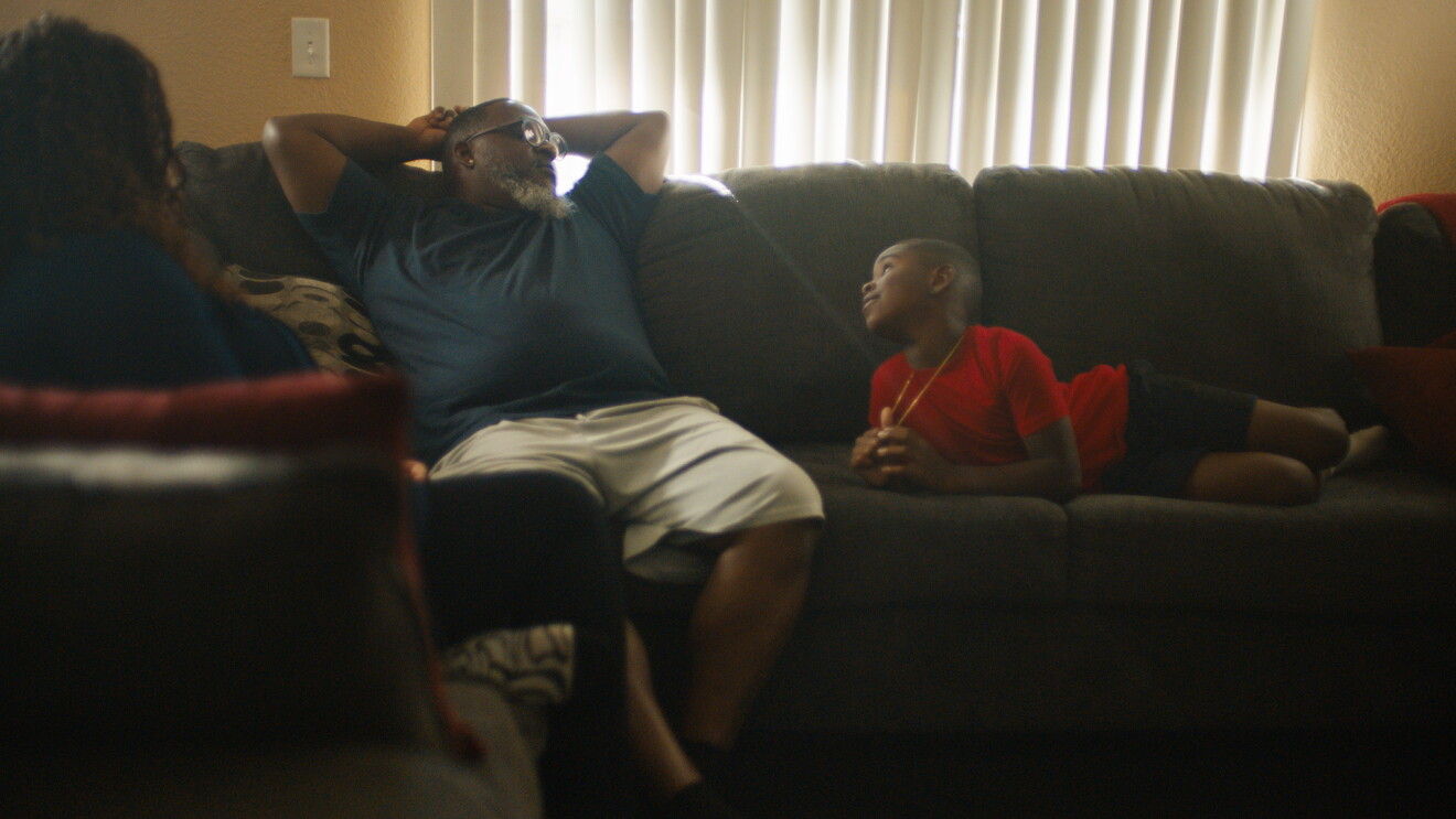 An image of a man and his son relaxing on a couch at home. The mother is also in the photo, sitting on the couch as well with her back to the camera.
