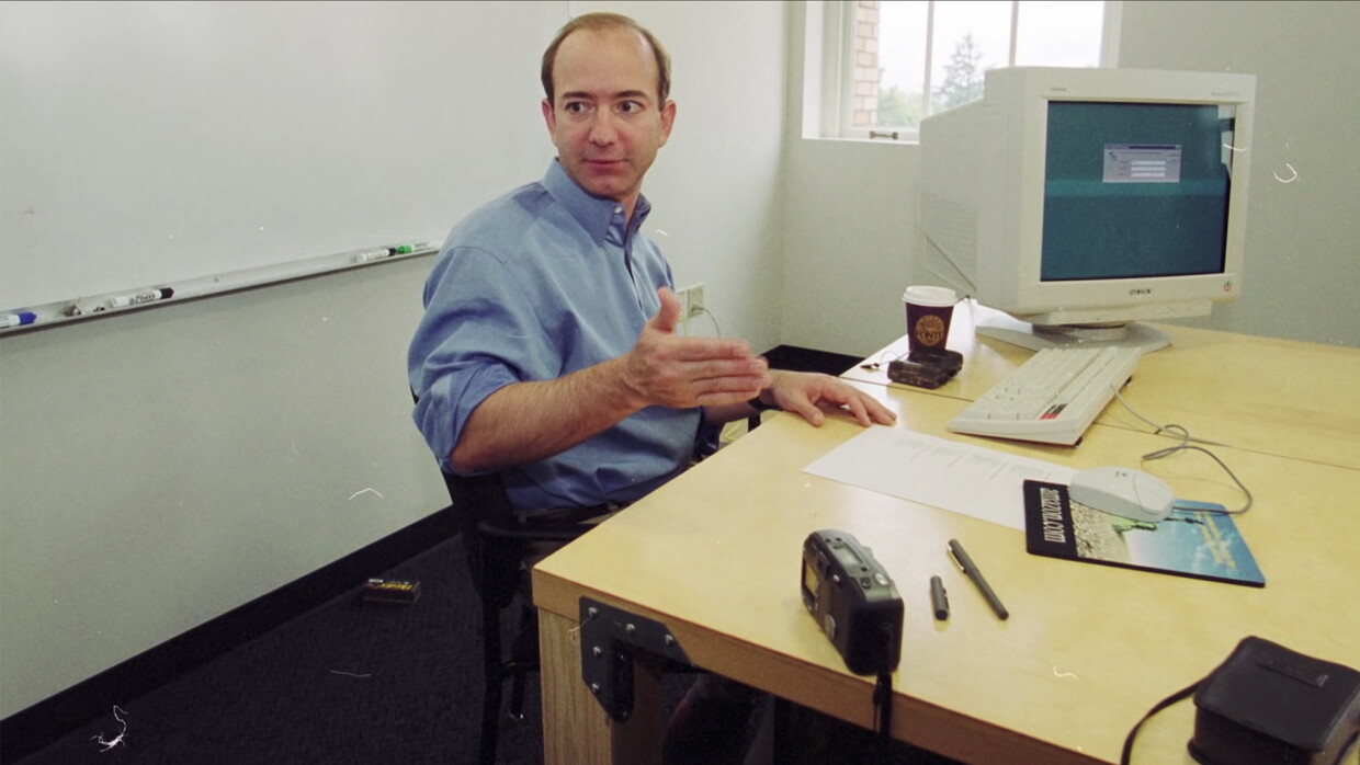 Jeff Bezos, muy joven, en su habitación de trabajo. De fondo una ventana y una pizarra blanca con los rotuladores de colores. Bezos está sentado en una silla negra y delante de una mesa de color madera. En la mesa hay un ordenador de mesa antiguo, un teclado y un ratón con su alfombrilla. En la mesa también un café con vaso de cartón, una cámara, un bolígrafo, un tapón de bolígrafo, una papel y una grabadora.
