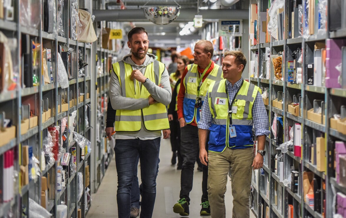 Christoph Metzelder läuft mit Mitarbeitern von  Amazon durch einen Regalgang des Logistikzentrums.