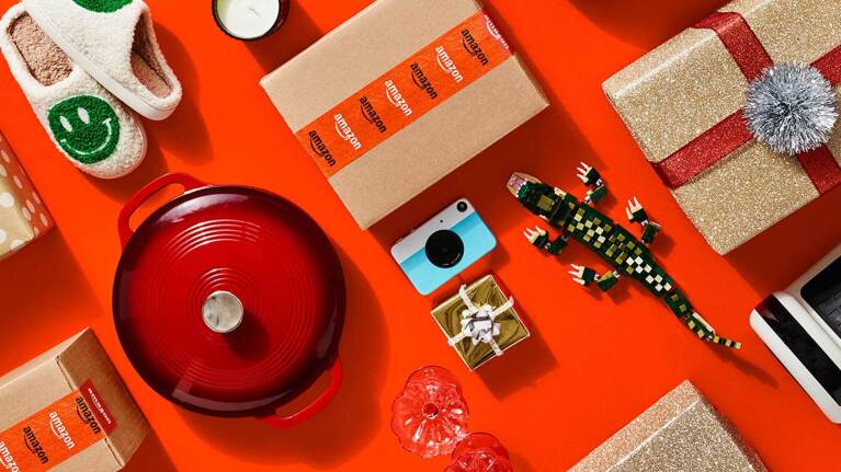 A collection of shipping boxes with Amazon labels, gift-wrapped presents, and unwrapped gifts decoratively placed on a red background.