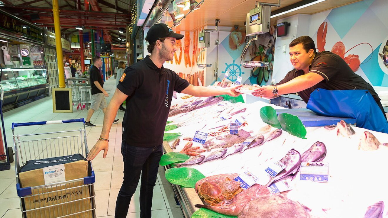 Mercado de La Paz de Madrid. Trabajador de Prime Now recoge en una pescadería el paquete de un cliente. El pescadero, con un delantal azul, tiene que incorporarse para entregarle el pedido. El trabajador de Prime Now va con un carro de la compra que contiene más pedidos. 
