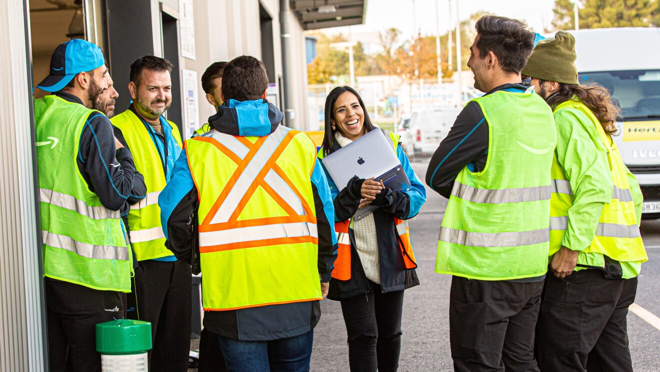 Ivette Duran, CEO de Cainaima Logistics, de pie con un ordenador entre las manos y hablando con su equipo de conductores de las furgonetas de reparto de paquetes. Están rodeada de 7 conductores y ella conversa con ellos con una sonrisa Ella es morena y tiene el pelo largo y negro. Lleva un jersei de color blanco y de cuello alto, una chaqueta azul y un chaleco fluorescente. Ellos la rodean y llevan todos pantalones negros y chalecos fluorescentes. De fondo la pared del centro logístico con una furgoneta.