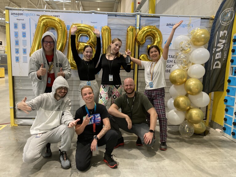 Mitarbeitende aus ganz Baden-Württemberg nahmen an der Initiative „Amazon Goes Gold for Kids with Cancer“ teil und posierten vor Wand mit weißen und goldenen Luftballons.